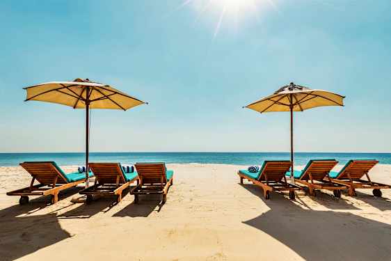 Liggestole med parasoller på en sandstrand, med havet i baggrunden. (AI-genereret tekst)