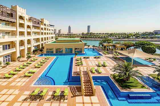 Et hotelområde med en stor pool, liggestole og grønne udendørsområder. Bygningen har flere etager og ligger ved vandet. (AI-genereret tekst)