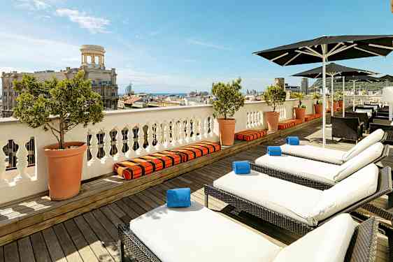 Udsigt over en terrasse med liggestole og parasoller, med en by og havet i baggrunden. (AI-genereret tekst)