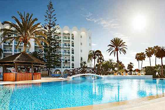 Billede af en feriested med en udendørs pool, en træbro over poolen, palmer og en hvid bygning i baggrunden. (AI-genereret tekst)