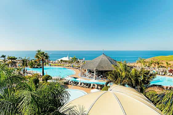 Et resortområde ved kysten med pooler, en pavillon og palmer, med havet i baggrunden. (AI-genereret tekst)