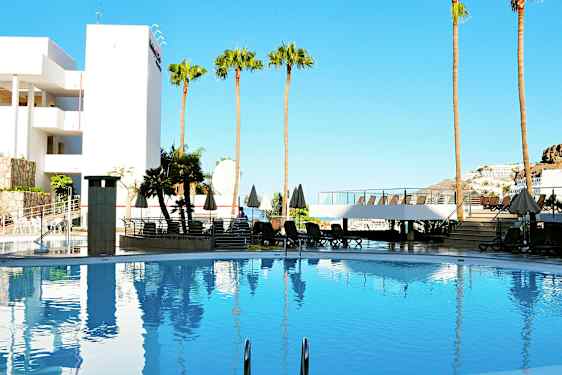 En swimmingpool omgivet af liggestole, parasoller og palmer, med en bygning og klar blå himmel i baggrunden. (AI-genereret tekst)