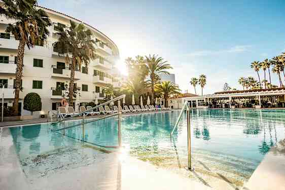 En udendørs pool foran et hotel omgivet af palmer og liggestole. (AI-genereret tekst)