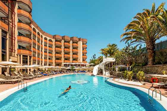 En pool med en kvinde der svømmer, omgivet af liggestole og et hotel i baggrunden. (AI-genereret tekst)