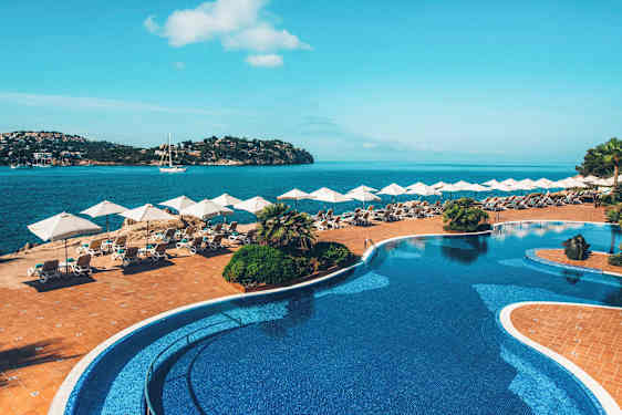 En pool med blå fliser ligger ved siden af en terrasse med solsenge og parasoller, med udsigt over en havbugt. (AI-genereret tekst)