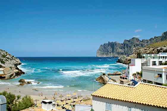 En smuk kyst med turkisblåt hav, sandstrand og parasoller. Der er klipper og bygninger med lyse tage. (AI-genereret tekst)