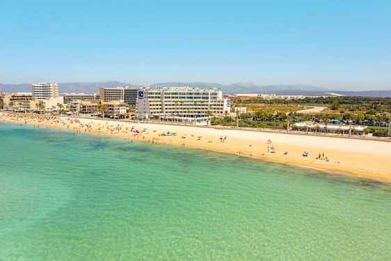 Sandstrand med badende personer og klart turkis vand. Hoteller og bygninger i baggrunden. (AI-genereret tekst)