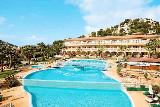 Billedet viser et hotel med en stor pool foran og en omkringliggende terrasse med liggestole. Baggrunden har en frodig bakke og klar blå himmel. (AI-genereret tekst)