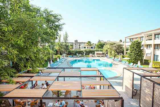 En ferieresort med en stor oval pool omgivet af liggestole og parasoller samt et spiseområde med pergolaer. (AI-genereret tekst)