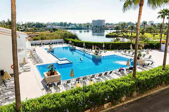 Udendørs swimmingpool med liggestole og parasoller omkring, omgivet af grønne planter og med udsigt over en sø og et åbent landskab. (AI-genereret tekst)