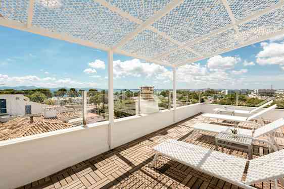 En tagterrasse med liggestole og et bord. Terrassen har et perforeret overdække og glasværn med udsigt over byen og havet. (AI-genereret tekst)