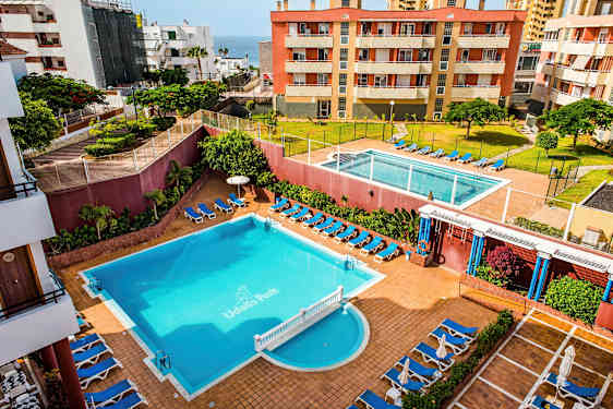 Et hotelområde med to swimmingpools, liggestole og omkringliggende bygninger. (AI-genereret tekst)