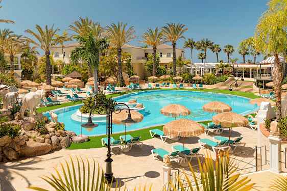 Et billede af et luksuriøst feriested med en pool, omgivet af liggestole og parasoller, omringet af palmer og bygninger. (AI-genereret tekst)