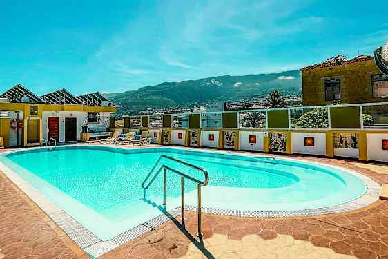 Udendørs swimmingpool med liggestole i en solrig atmosfære, omgivet af bygninger og med bjerge i baggrunden. (AI-genereret tekst)