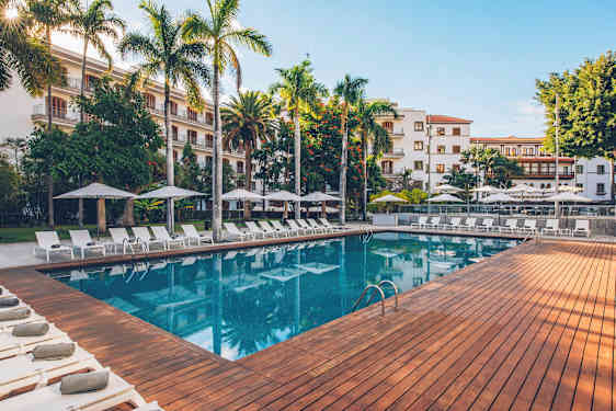 En udendørs pool omgivet af liggestole, parasoller og frodige træer, med en bygning i baggrunden. (AI-genereret tekst)