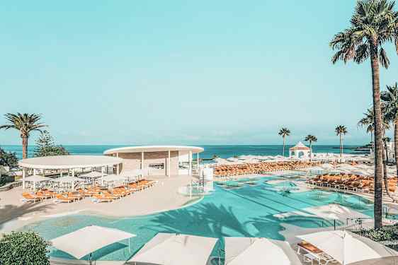 Et poolområde med parasoller og liggestole, omgivet af palmer og med havudsigt. (AI-genereret tekst)