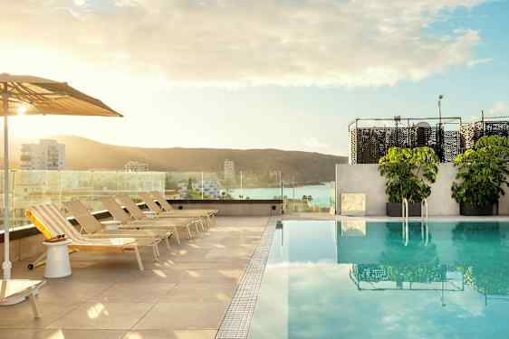Udsigt over en pool med liggestole og parasoller, i baggrunden ses en by og havet. (AI-genereret tekst)