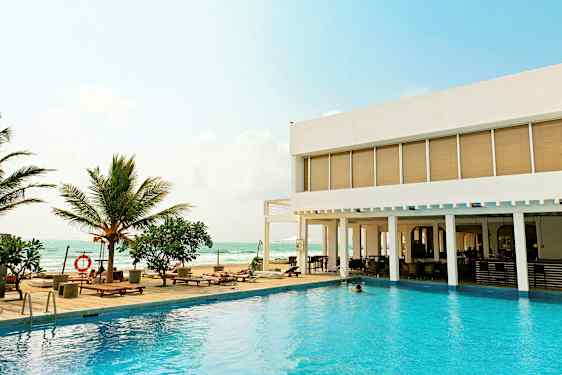 En udendørs pool med liggestole og palmer ved siden af et moderne hotel med udsigt over havet. (AI-genereret tekst)