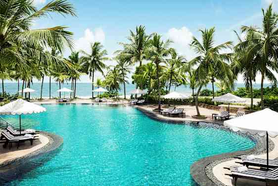 Billede af en swimmingpool omgivet af palmer og liggestole med parasoller ved havet. (AI-genereret tekst)