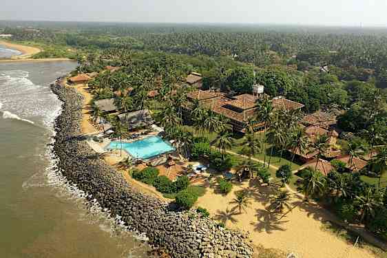 Udsigt over en kystlinje med et hotelområde, der inkluderer swimmingpool, bygninger med tegltage og omgivelser med frodig vegetation og kokospalmer. (AI-genereret tekst)