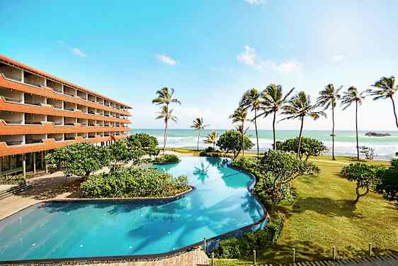 En hotelbygning ved stranden med en stor pool, palmer og havet i baggrunden. (AI-genereret tekst)