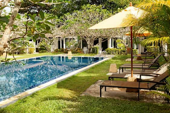 En swimmingpool med grønne områder og liggestole ved siden af, samt en række hvide bygninger i baggrunden. (AI-genereret tekst)