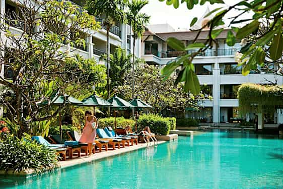 Billede af en udendørs swimmingpool omgivet af liggestole, parasoller og planter, med en bygning i baggrunden med altaner. (AI-genereret tekst)