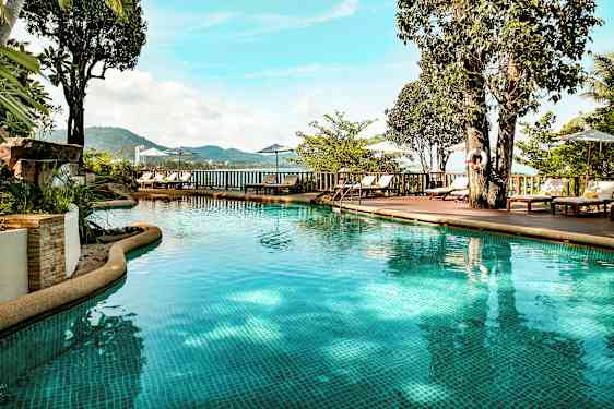 Udendørs pool med klart vand omgivet af liggestole, parasoller og grønt. Udsigten viser bjerge og hav i baggrunden. (AI-genereret tekst)