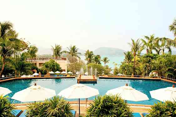 Billede af et feriested med en udendørs pool omgivet af parasoller og palmer, med udsigt til havet og bjerge i baggrunden. (AI-genereret tekst)