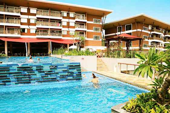 Et hotel med to etager er synligt bag en udendørs pool, hvor mennesker svømmer. Grønne planter og terrasseområder omkranser poolområdet. (AI-genereret tekst)