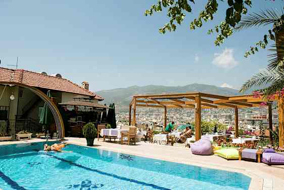 En udendørs pool ved et hotel med solsenge og parasoller. En træpergola og udsigt over en by og bjerge i baggrunden. (AI-genereret tekst)