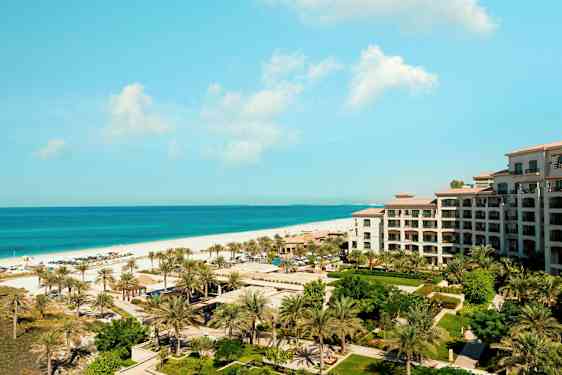 Udsigt over et feriested ved havet med en bygning, grønne områder og palmer i forgrunden. (AI-genereret tekst)