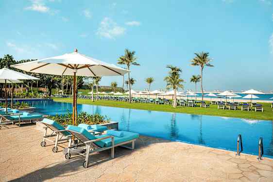 En pool med liggestole og parasoller i et udendørsmiljø, omgivet af palmer og græsplæne. (AI-genereret tekst)