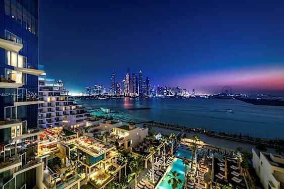 Udsigt over et moderne bymiljø ved skumring med skyskrabere, hav og en bygning med oplyste terrasser og poolområde i forgrunden. (AI-genereret tekst)