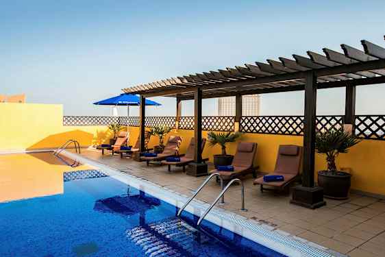 En udendørs pool med blåt vand omgivet af solvogne og parasoller. Solvognene står på et gulv med fliser med en gul væg og en træpergola i baggrunden. (AI-genereret tekst)