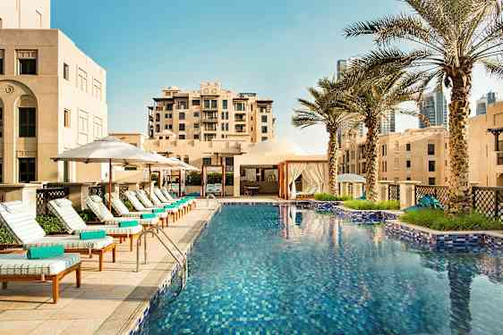 En udendørs pool omgivet af liggestole med parasoller og palmer ved siden af bygninger. (AI-genereret tekst)