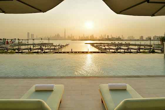 Udsigt fra en infinitypool mod en marina og en bysilhuet under en solnedgang, med to liggestole i forgrunden. (AI-genereret tekst)