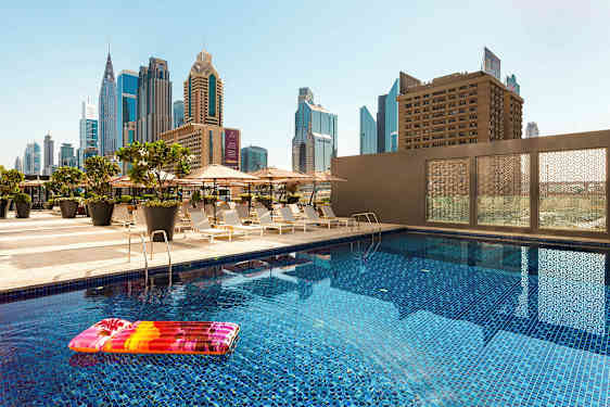 En udendørs pool med en oppustelig strandmadrasse, omgivet af liggestole og parasoller. Høje bygninger kan ses i baggrunden. (AI-genereret tekst)