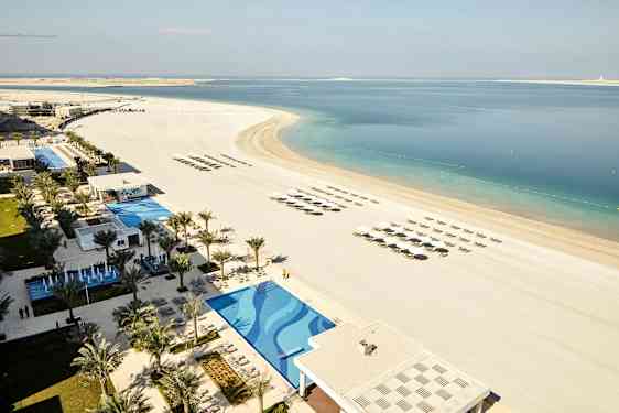 Billedet viser en strand med parasoller og solsenge i lige rækker, ved siden af en pool og et resortområde omgivet af palmer. (AI-genereret tekst)