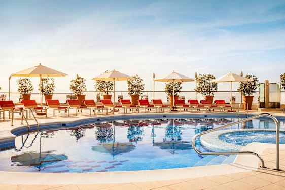 En udendørs swimmingpool omgivet af røde liggestole, hvide parasoller og potteplanter. (AI-genereret tekst)