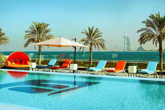 En pool med liggestole og parasoller, omgivet af palmer med havudsigt og en skyline i baggrunden. (AI-genereret tekst)