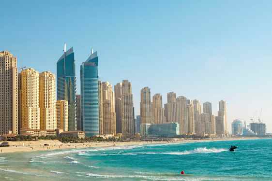 Billede af højhuse ved en strand med klart blåt vand, en person er på en jetski nær stranden. (AI-genereret tekst)