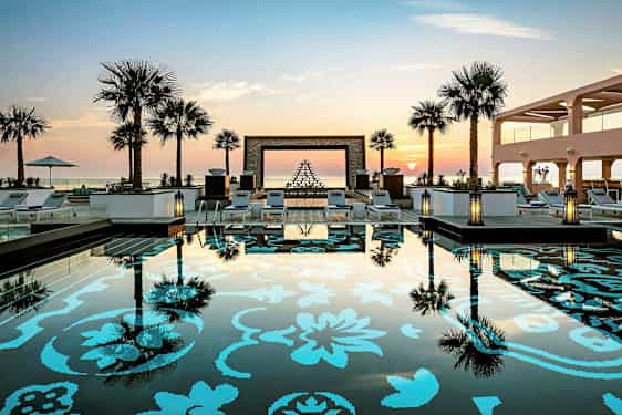 Billede af en pool med dekorativt mønster og omgivelser med solsenge, palmer og en solnedgang i baggrunden. (AI-genereret tekst)