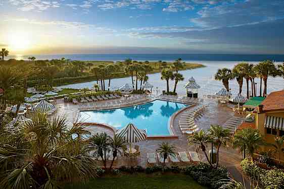 Billede af et poolområde med liggestole og parasoller, omgivet af palmer og en strand i baggrunden. (AI-genereret tekst)