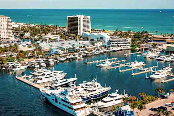 En lystbådehavn med luksusyachter og både ved siden af urbane bygninger og grønne områder, med havet i baggrunden. (AI-genereret tekst)