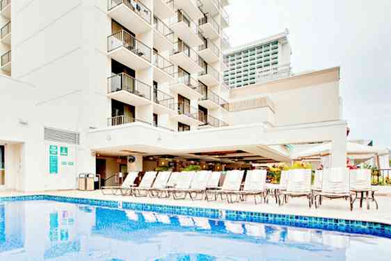 Billede af en bygning med altaner, en pool og liggestole. (AI-genereret tekst)