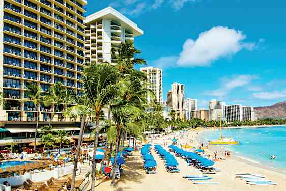 En sandstrand med liggestole og blå parasoller, omgivet af høje hotelbygninger og palmer. (AI-genereret tekst)