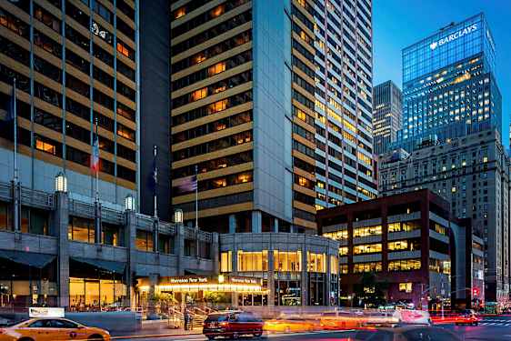 Et billede af et bymiljø med høje bygninger, hvoraf en bygning har teksten "Sheraton New York" ved indgangen. Taxaer er synlige på gaden. (AI-genereret tekst)
