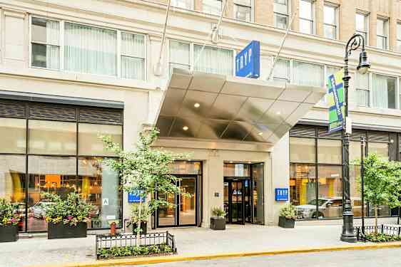 Indgangen til et hotel med skiltet "TRYP". Bygningen har store vinduer og plantekasser. (AI-genereret tekst)