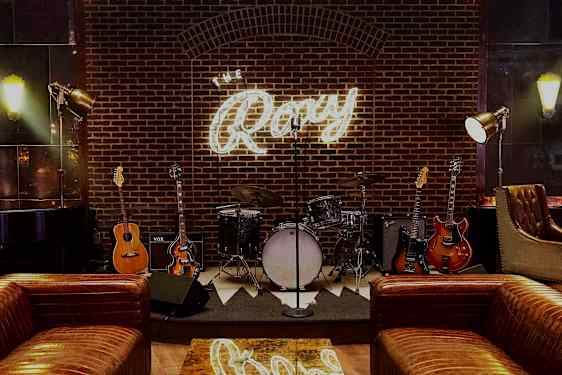 En scene med musikinstrumenter som trommer og guitarer foran en murstensvæg med et lysende skilt, der siger "The Roxy". (AI-genereret tekst)